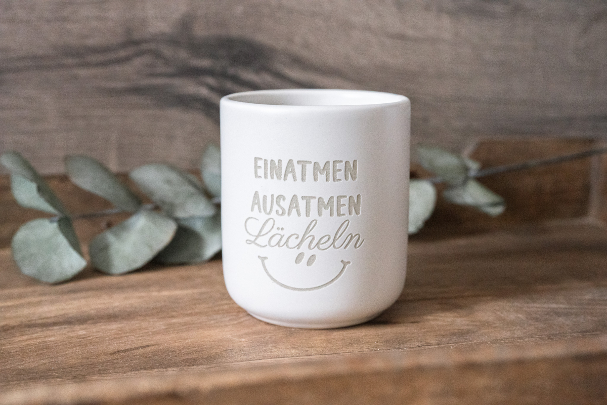 Matte weiße Keramiktasse mit Gravur „EINATMEN AUSATMEN Lächeln“ und Smiley auf Holzuntergrund, Eukalyptus im Hintergrund.